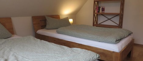 Ein Zimmer mit zwei Holzbettgestellen, jeweils mit einem Kopfkissen und einer Decke. Gegen die Wand steht ein Bücherregal aus Holz.