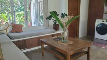 A room with a wooden bench, a small table with a vase of flowers, a washing machine, and a window with curtains.