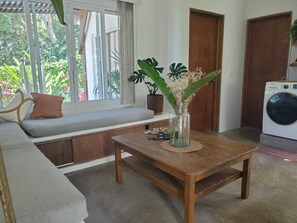 Une pièce avec un banc en bois, une petite table sur laquelle est posé un vase de fleurs, un lave-linge et une fenêtre avec des rideaux.