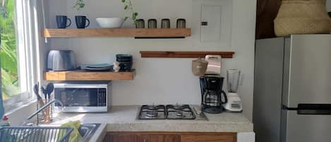 A kitchen with wooden shelves, a gas stove, a microwave, a coffee maker, and a refrigerator.