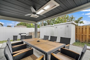 Un patio exterior con mesa y sillas, un ventilador de techo y una parrilla.