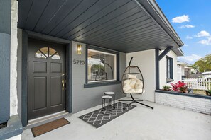 Ein Wohnhaus mit dunkler Haustür, einer Hausnummer und einem Hängekorbsitz sowie einem kleinen Tisch.