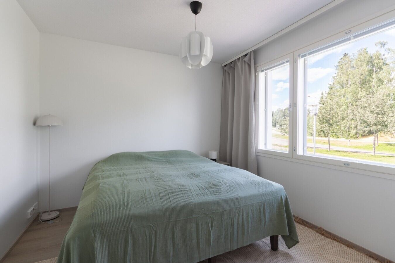 Een slaapkamer met een bed, een vloerlamp, een raam met gordijnen en uitzicht op bomen.