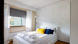Una habitación de hotel con pared de ladrillo blanco, una cama con ropa blanca y almohadas de colores, una ventana con cortinas blancas y un radiador.