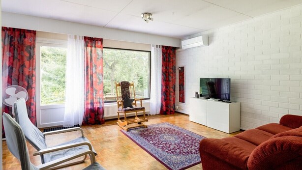 Una sala de estar con piso de madera, un sofá rojo, un televisor sobre un mueble blanco, un sillón mecededor y una ventana con cortinas rojas.