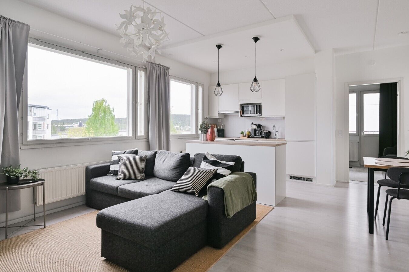 A modern living room with a grey sofa, a dining area with a table and chairs, and a kitchen with white cabinets and a microwave.