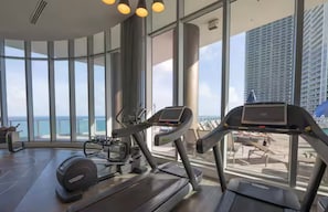 Ein Fitnessraum mit Laufbändern und Rudergerät, großen Fenstern und Blick auf das Meer sowie ein Hochhaus.