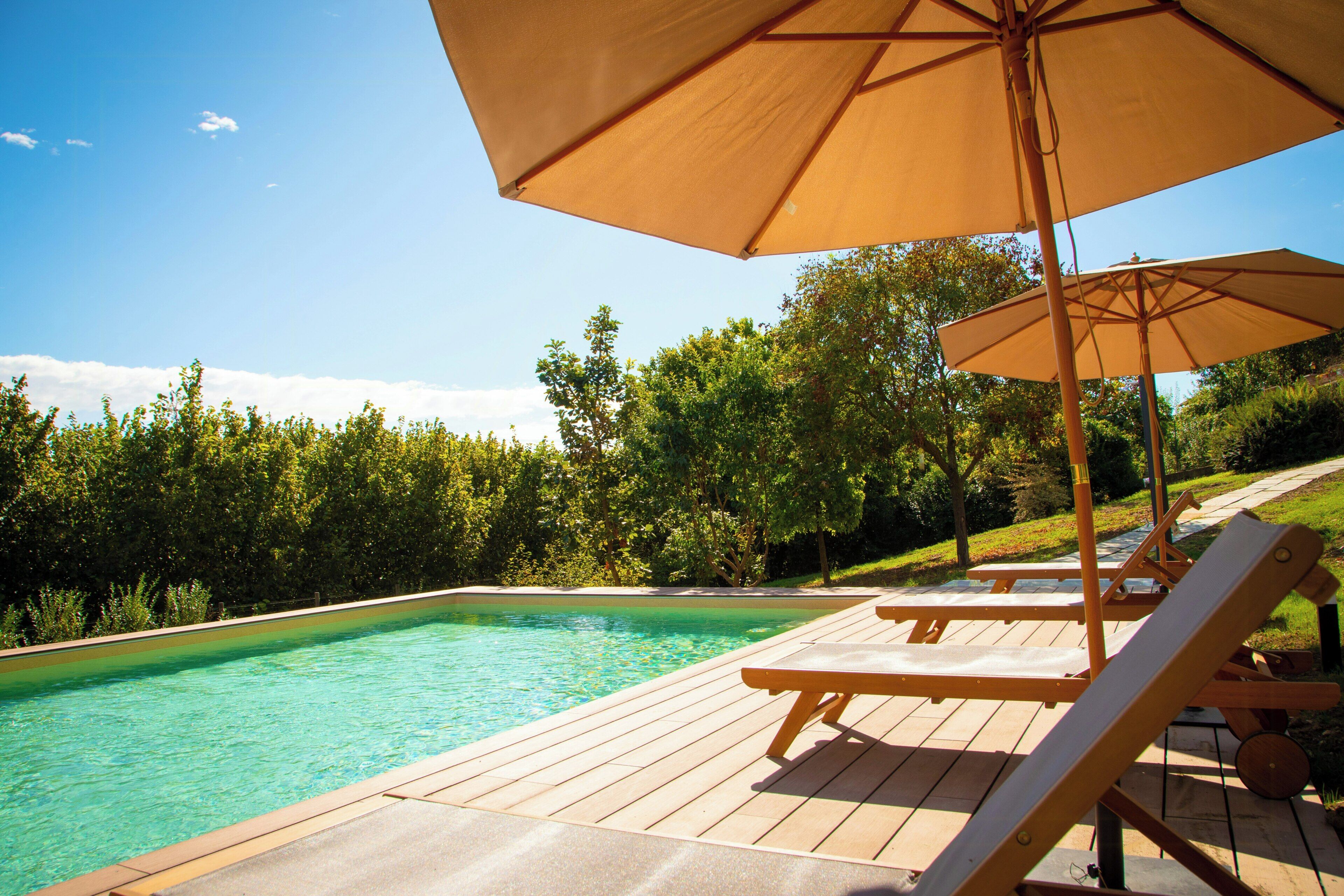 A swimming pool with wooden deck chairs and umbrellas, surrounded by lush greenery.
