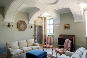 Ein Zimmer mit einem weißen Sofa, einem blauen Hocker, zwei gestreiften Sesseln, einem Kamin und einem Wappenbild an der Wand.