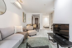 Ein modernes Wohnzimmer mit Sofa, Hocker, Couchtisch und Stühlen. Es gibt einen Essbereich mit Tisch und Stühlen sowie ein Schlafzimmer mit Bett. Der Raum hat weiße Wände und eine Decke mit Einbauleuchten.