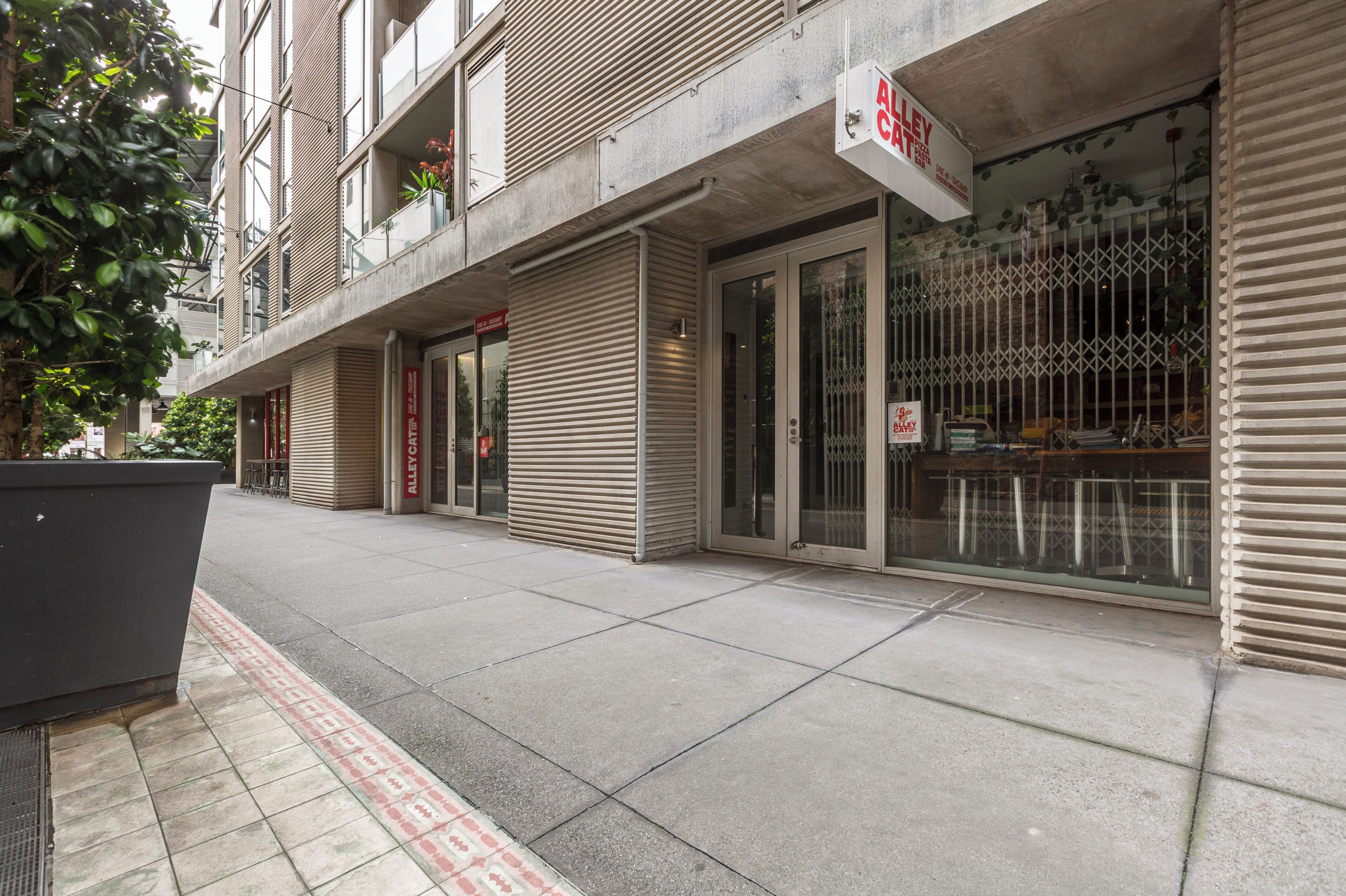 Vista da rua com lojas fechadas, incluindo uma pizzaria, e um edifício com entrada visível.