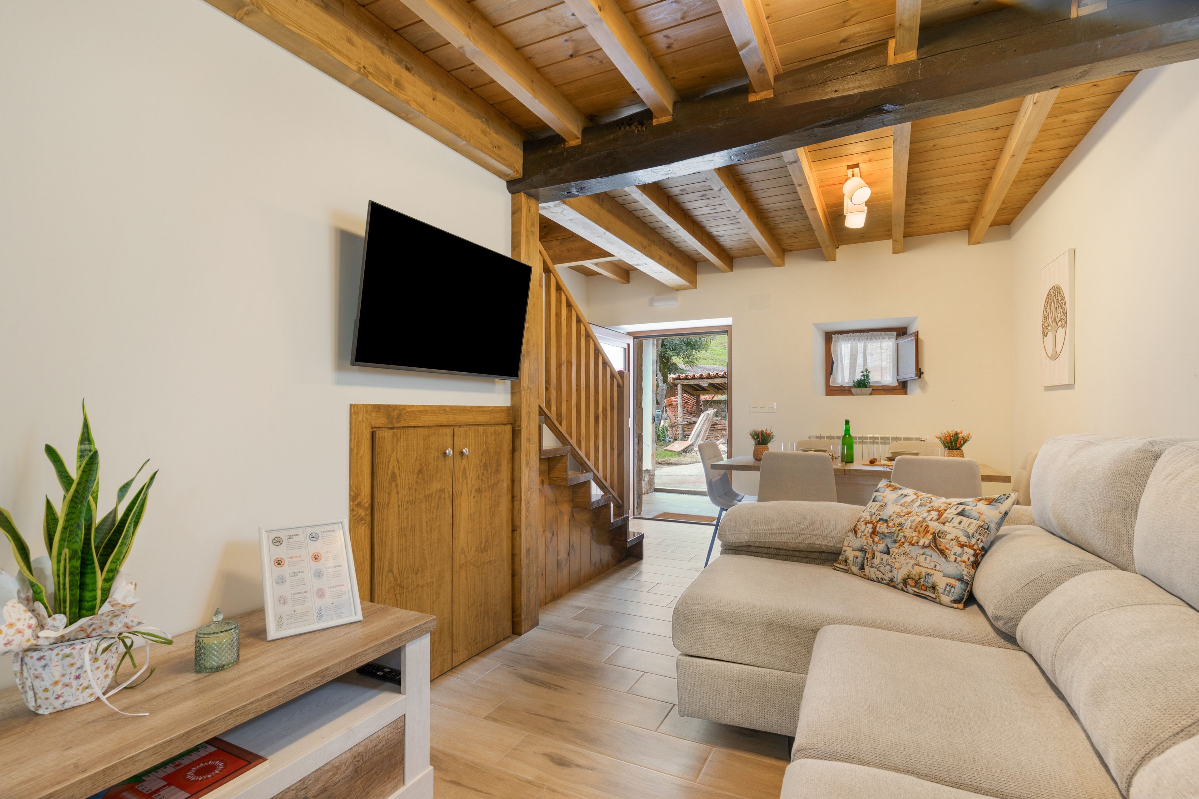Een moderne woonkamer met een houten plafond, een flatscreen televisie aan de muur, een bank met gekleurde kussens, en een houten kast met een menukaart in een lijst.