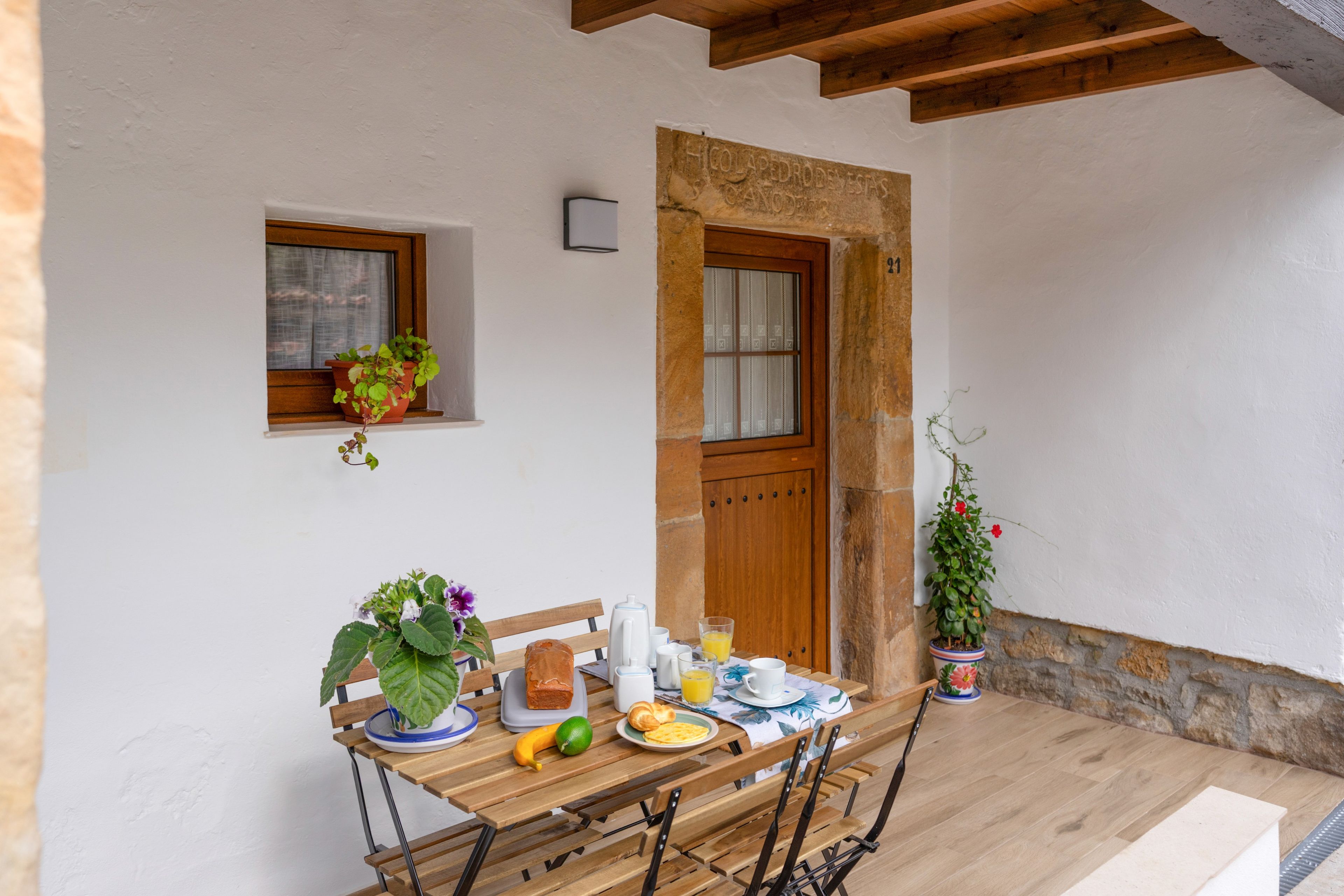 Una mesa de madera preparada para el desayuno, con una ventana, una puerta y plantas en macetas.