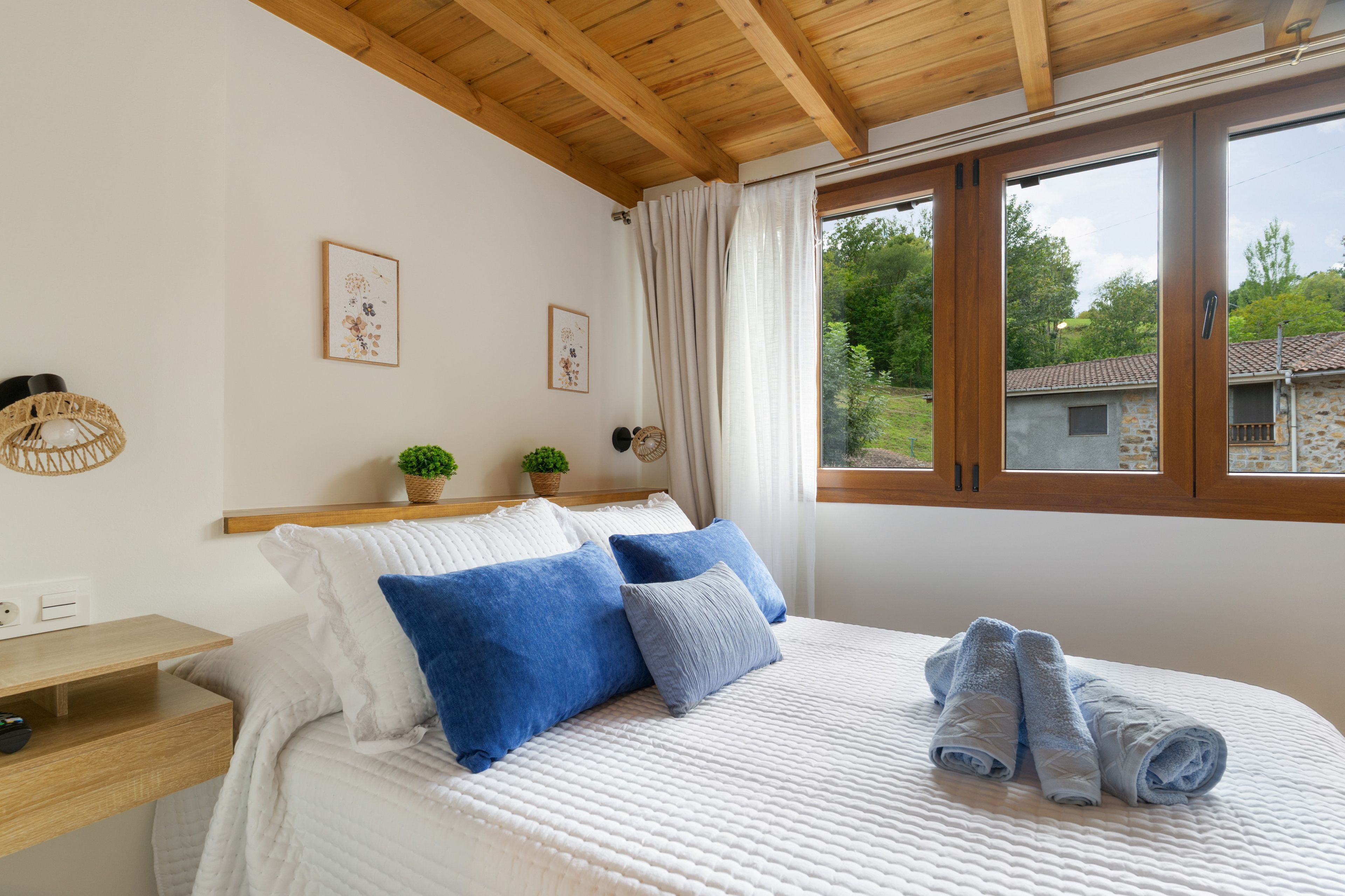 Un dormitorio con techo de madera, un ventanal con cortinas, una cama con ropa de cama blanca y almohadas azules, dos relojes en la pared y dos plantas en macetas.