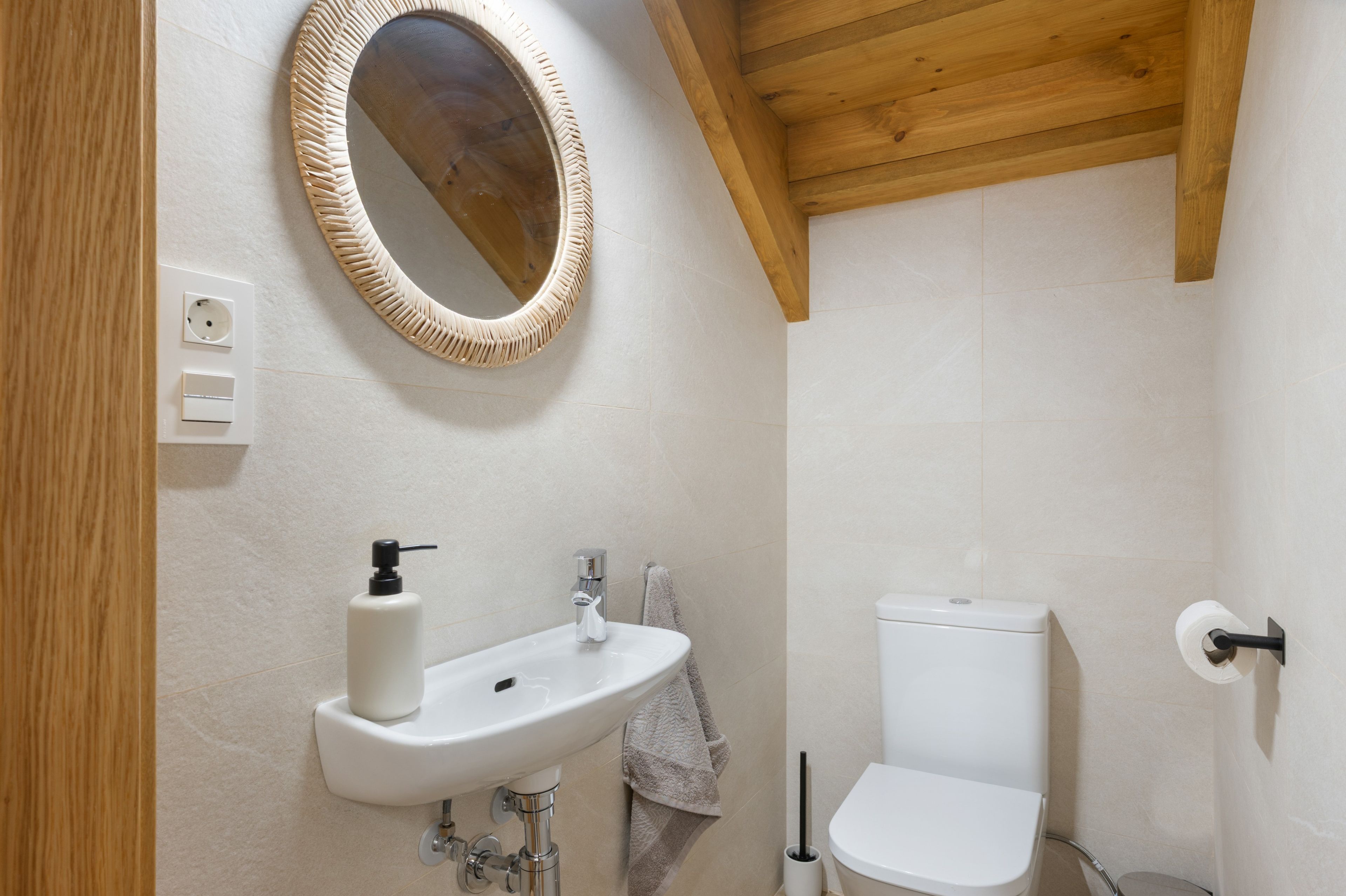 Een badkamer met een houten plafond, een ronde spiegel, een witte wasbak, een toilet en een handdoek aan de muur.