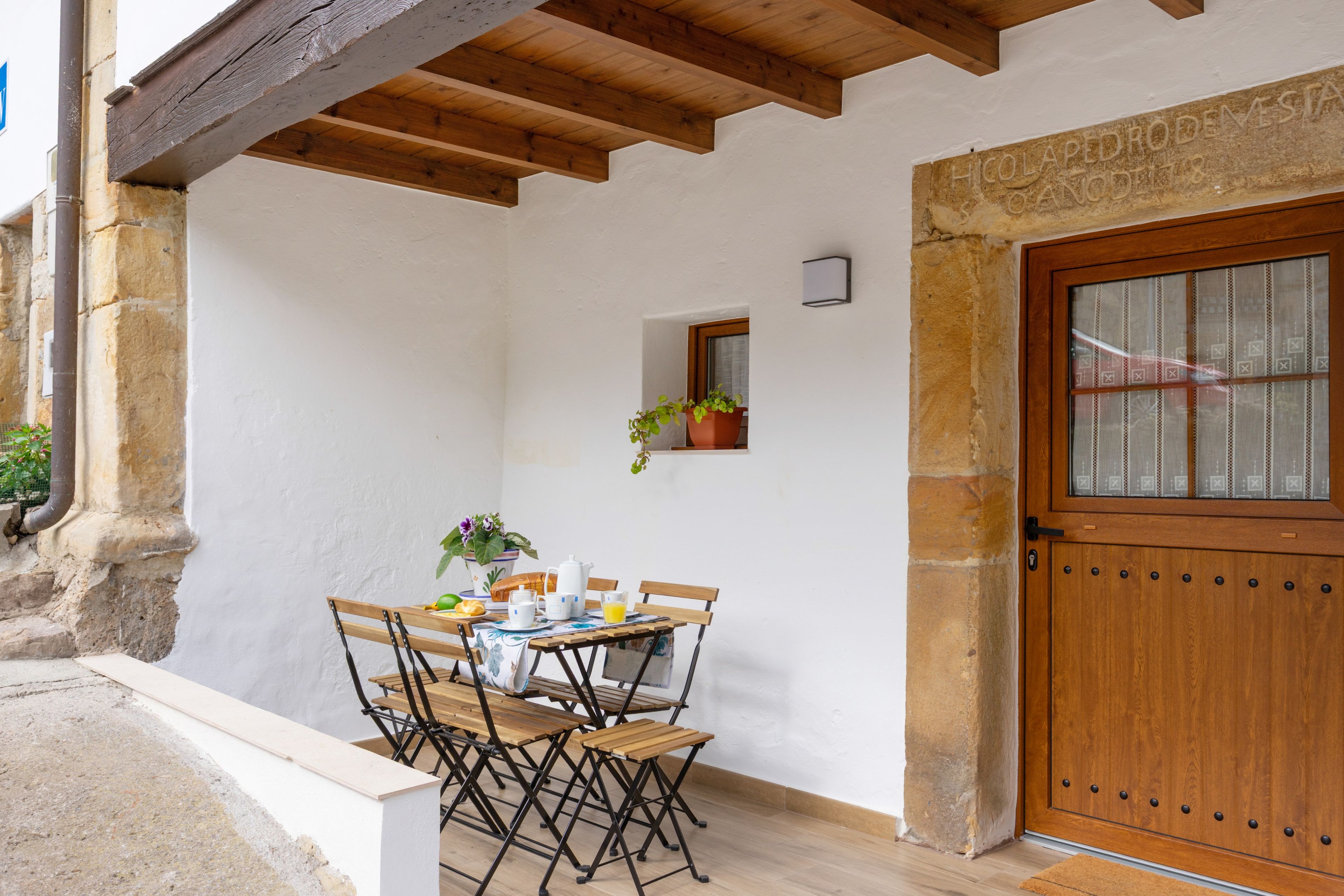Un coin repas avec une table et des chaises en bois, une plante en pot et une porte ornée d’une inscription gravée dans la pierre.