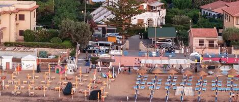 A coastal town with a beach area featuring rows of sun loungers and umbrellas, a small building with a sign, and residential buildings in the background.