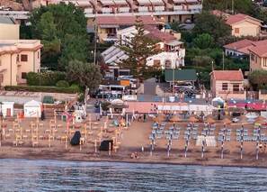 A coastal town with a beach area featuring rows of sun loungers and umbrellas, a small building with a sign, and residential buildings in the background. - Margherita-3 by Appartamenti Margherita seafront accommodation in Giglio Campese (Giglio Campese)