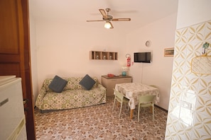 A small room with a patterned floor, a sofa with cushions, a table with chairs, a ceiling fan, a television, and a wall-mounted shelf. - Margherita-3 by Appartamenti Margherita seafront accommodation in Giglio Campese (Giglio Campese)