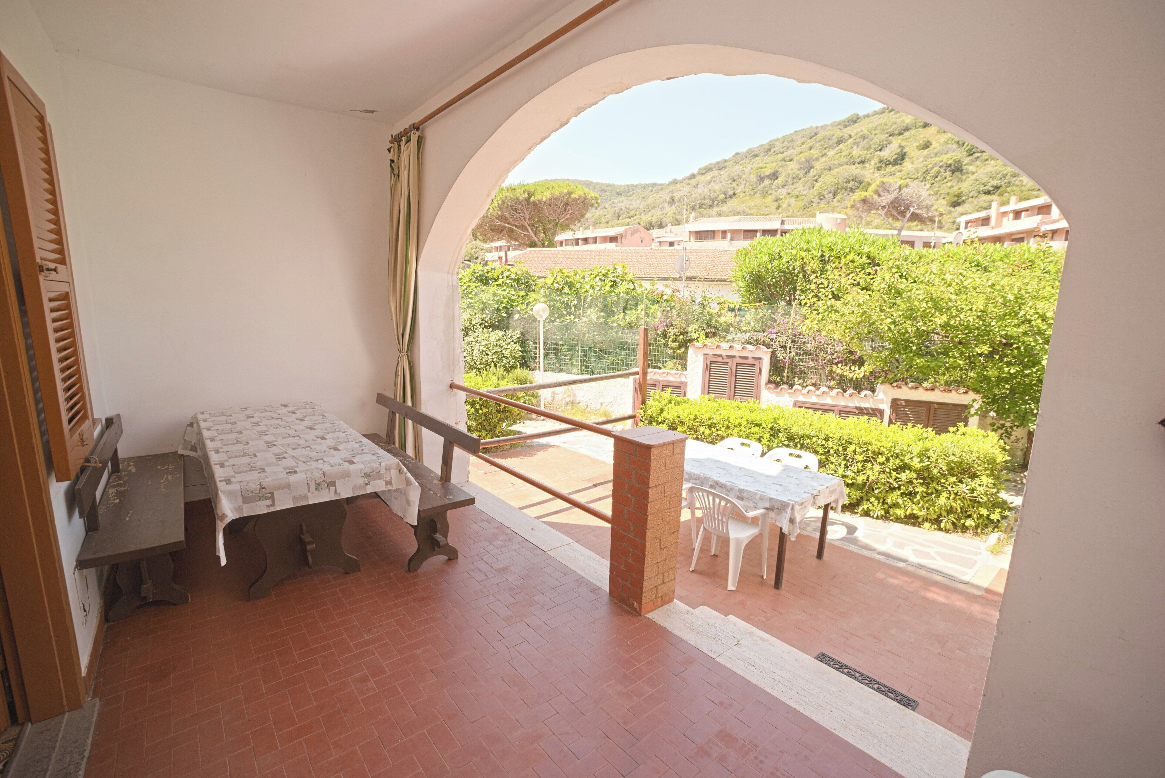A room with a table and chairs, an arched doorway leading to a balcony with a view of greenery and buildings.