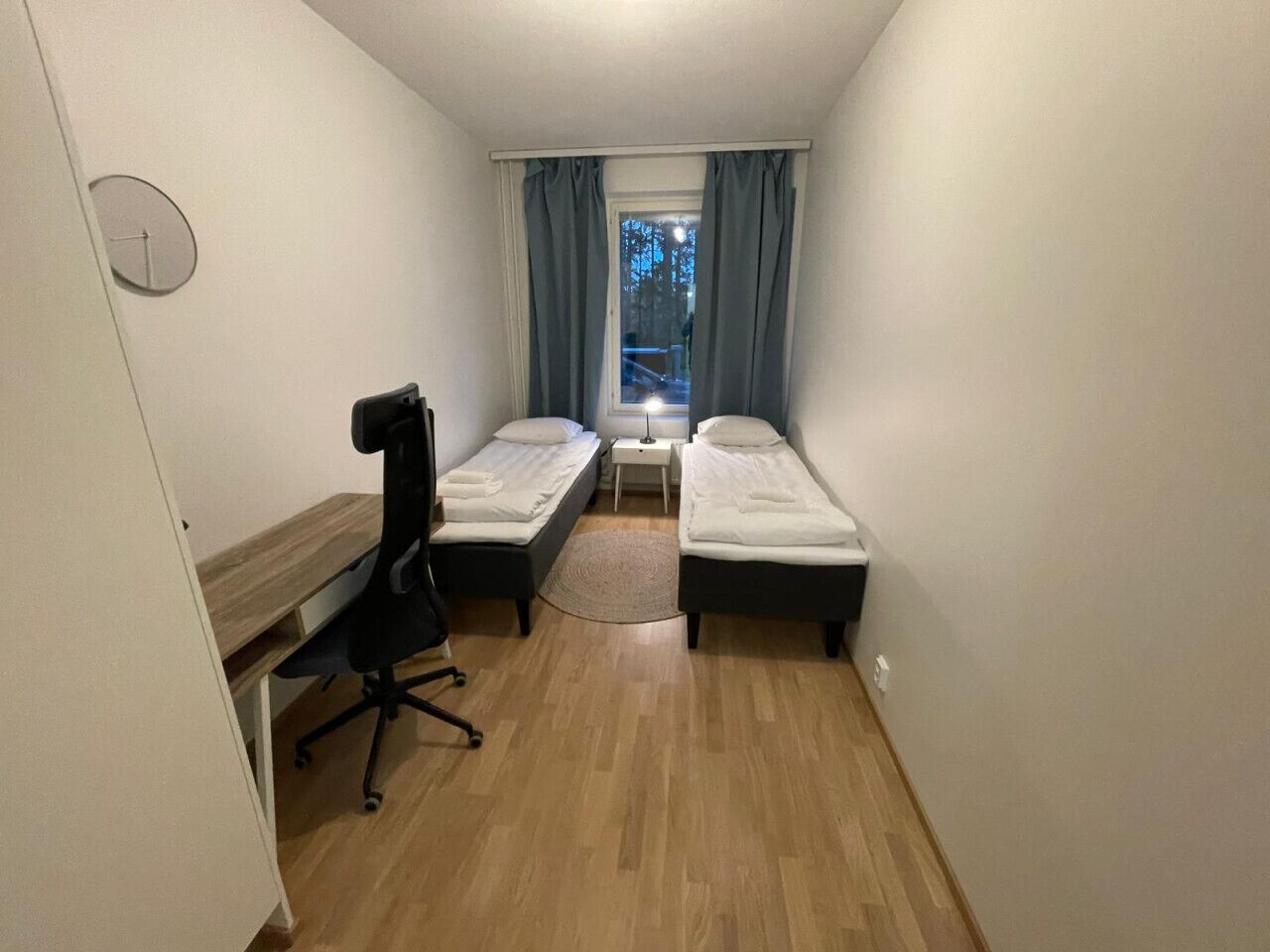 Una habitación pequeña con dos camas individuales, un escritorio, una silla y una ventana con cortinas.