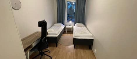 Una habitación pequeña con dos camas individuales, un escritorio, una silla y una ventana con cortinas.