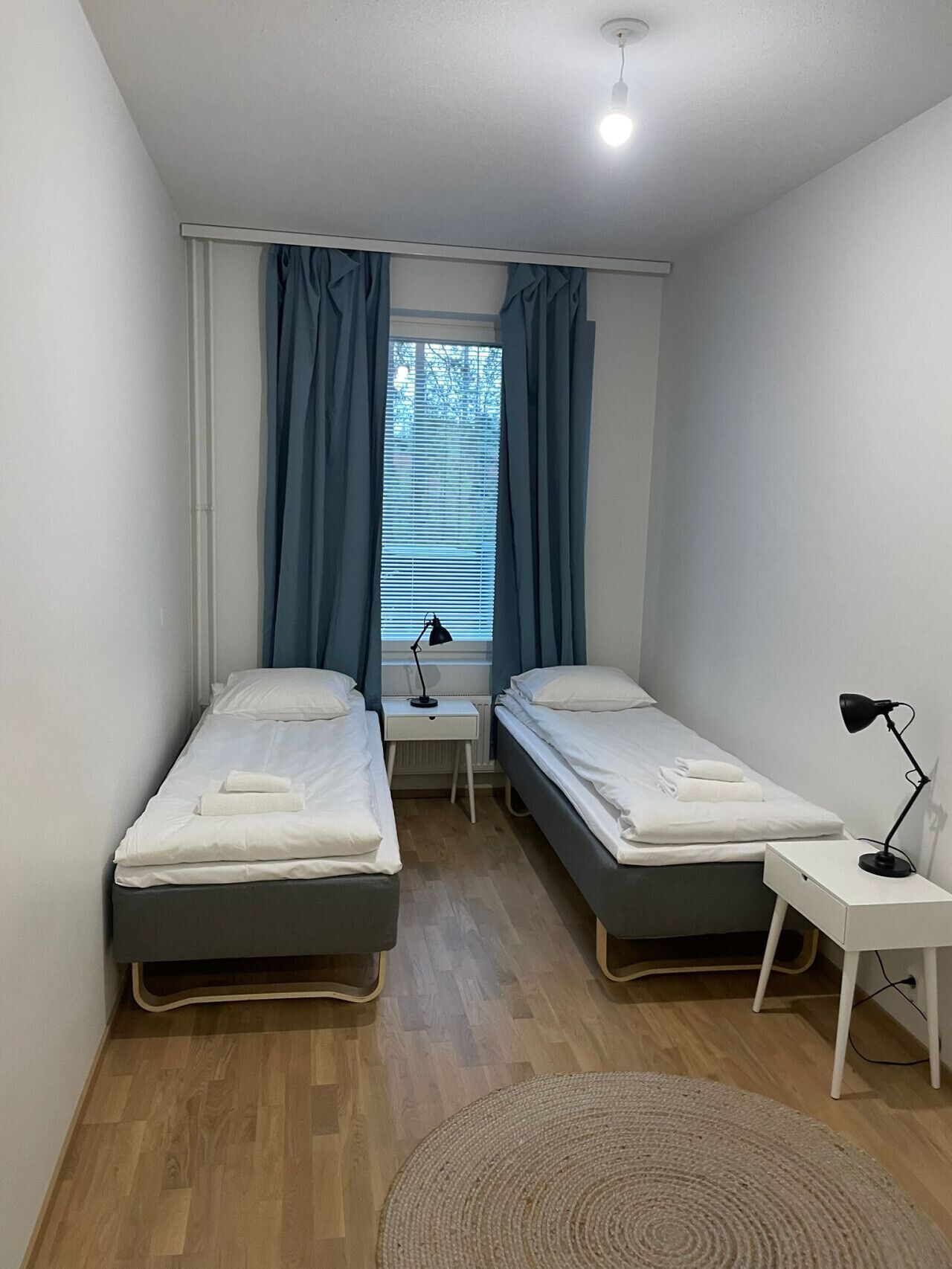 Pieni, siisti huone, jossa on kaksi erillistä sänkyä, ikkuna, jossa on verhot, yöpöytä lampun kanssa ja pyöreä matto lattialla.