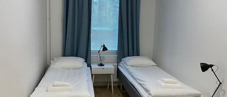 Pieni, siisti huone, jossa on kaksi erillistä sänkyä, ikkuna, jossa on verhot, yöpöytä lampun kanssa ja pyöreä matto lattialla.