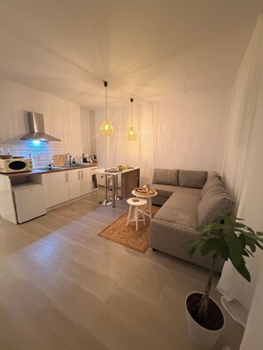A modern kitchen with white cabinets, a stainless steel range hood, and a microwave. There is a dining area with a small round table and a sofa. The room has a wooden floor and a large potted plant. - Apartment in Thiseio, just a stone's throw from the Acropolis (Athina)