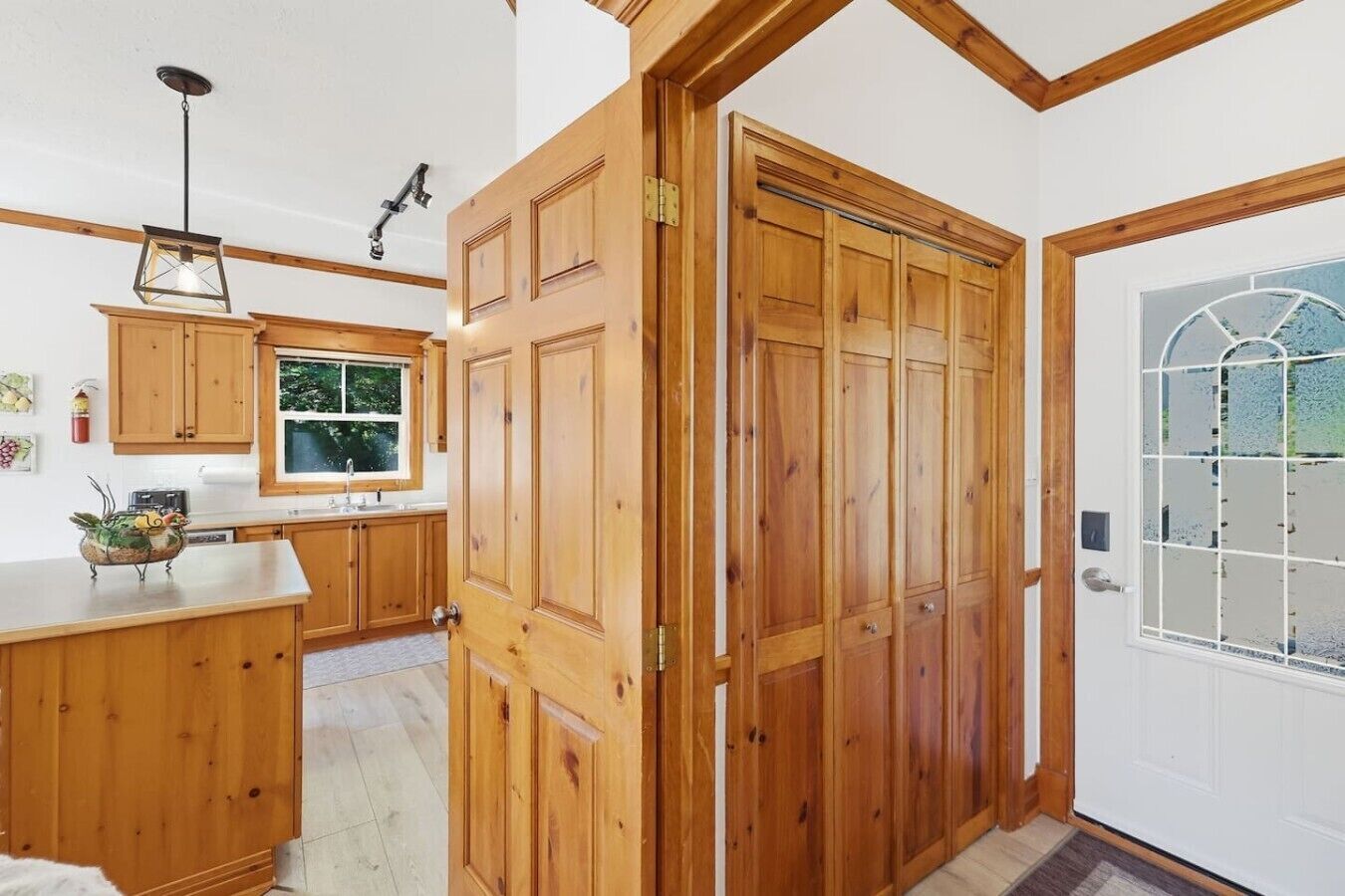 A wooden door with a glass panel, a kitchen with wooden cabinets and a window, and a hanging light fixture.