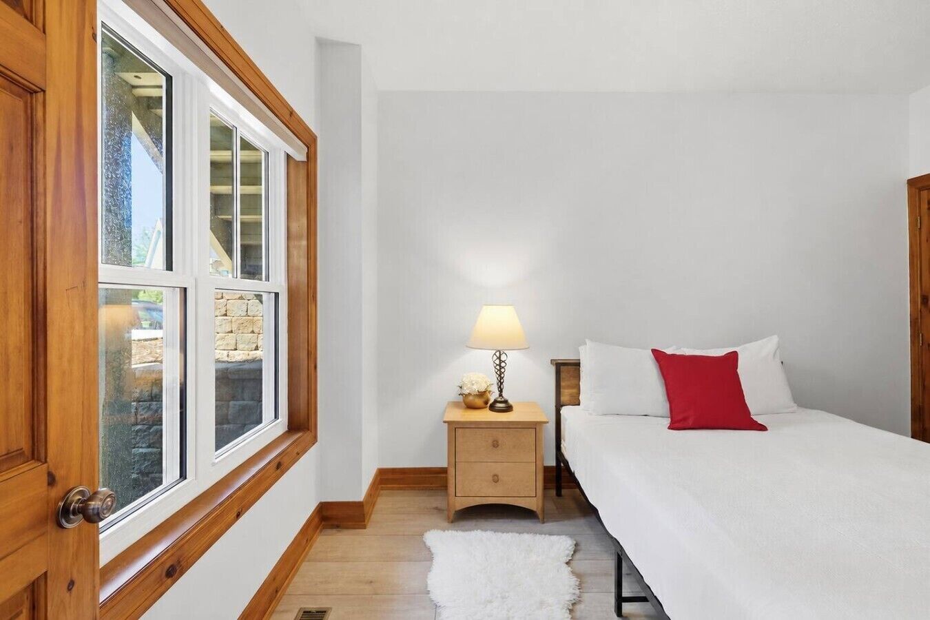 Un dormitorio con una cama, una mesita de noche con lámpara, una puerta de madera y una ventana con vista a un muro de piedra.