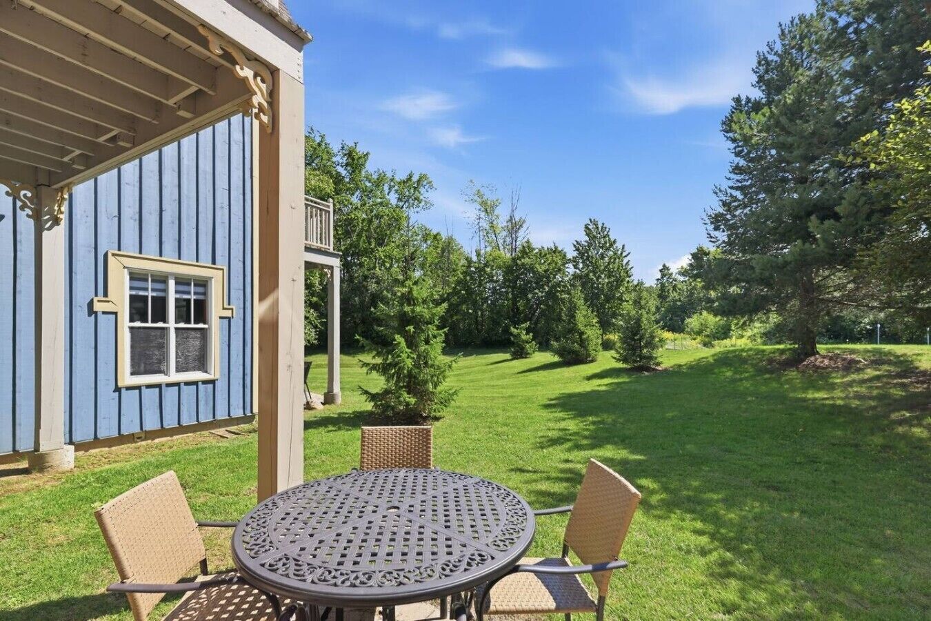 A blue building with a covered porch, a round metal table with chairs, and a grassy yard with trees.