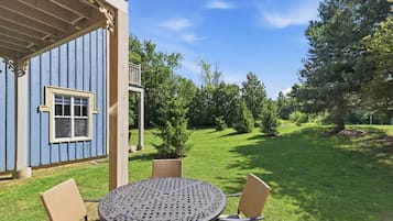 A blue building with a covered porch, a round metal table with chairs, and a grassy yard with trees.