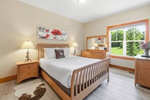 A bedroom with a wooden bed, bedside tables with lamps, a dresser, a mirror, and a window with a view of greenery. - Charming Snowbridge Condo with Summer Pool (The Blue Mountains)