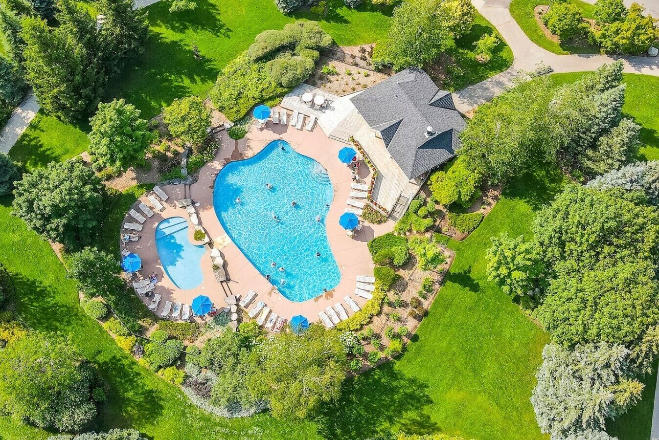 A large swimming pool surrounded by lounge chairs and umbrellas, with a covered patio area and a garden.