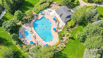 A large swimming pool surrounded by lounge chairs and umbrellas, with a covered patio area and a garden.