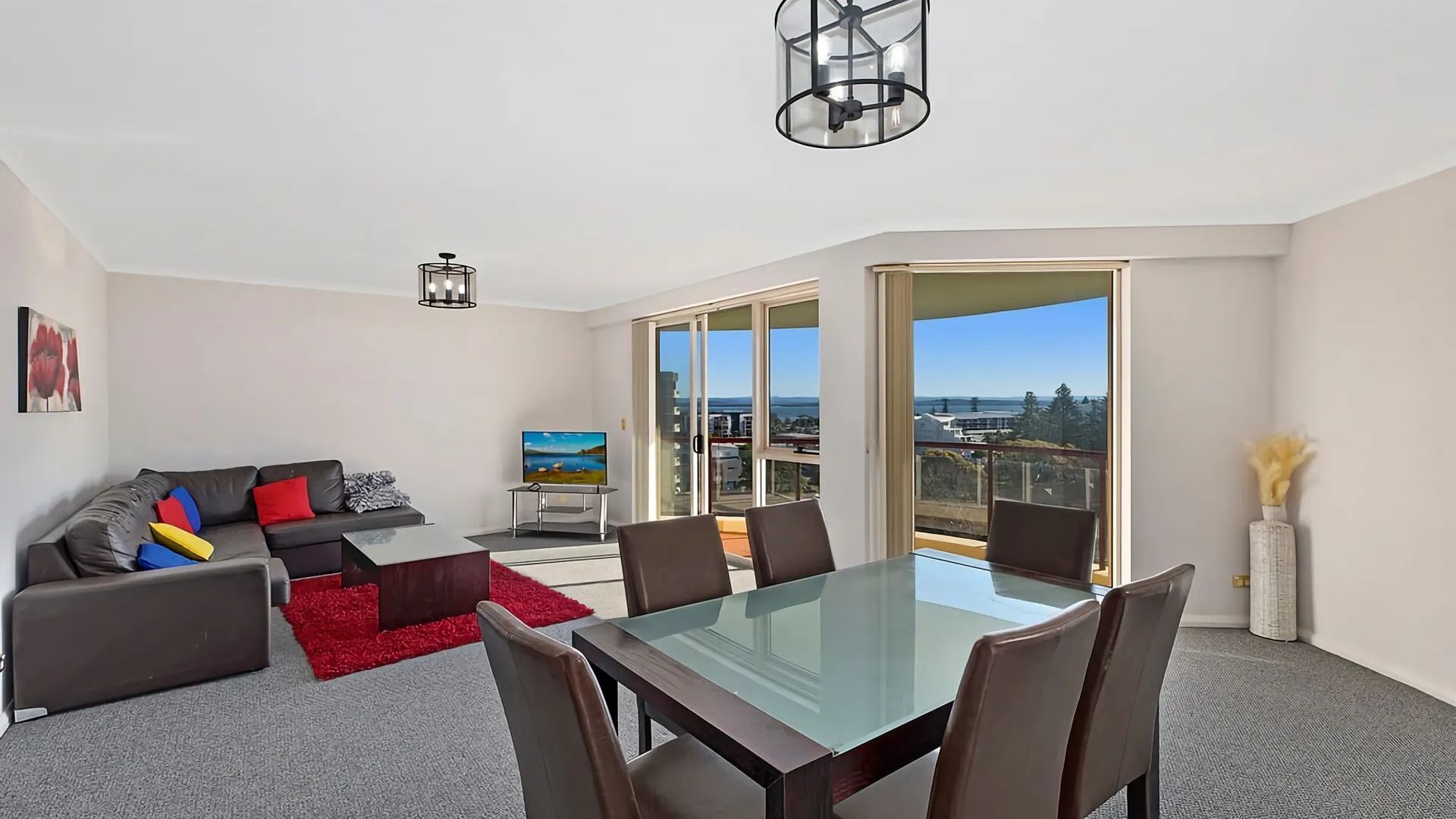 A modern living room with a glass dining table, a sofa set, and a large window offering a city view.
