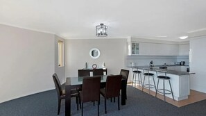 A modern kitchen with white cabinets, a central island with bar stools, and a dining area with a glass table and chairs. - Eagle Wings - Stunning Ocean Views - Unit 16 (The Entrance)