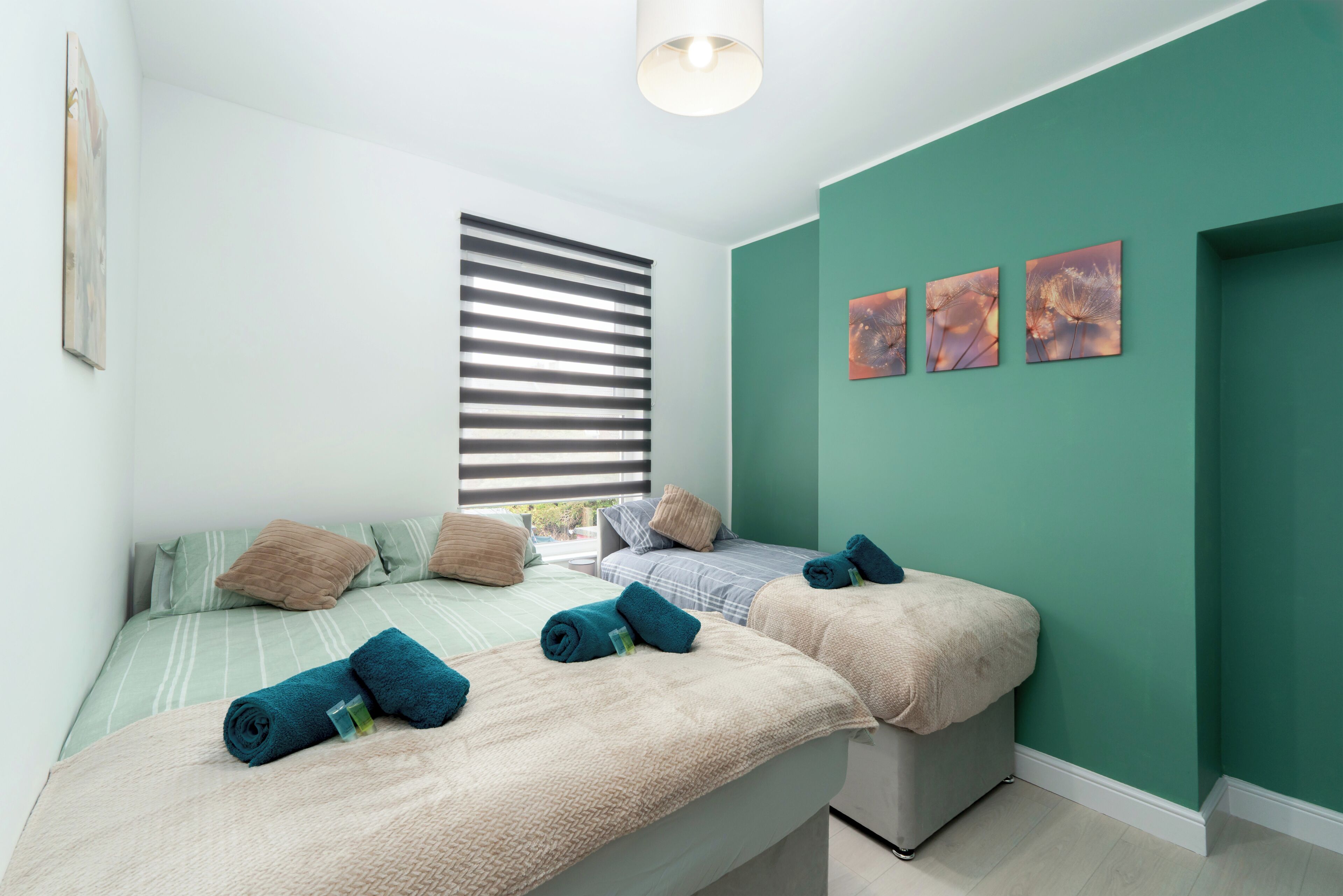 A bedroom with two beds, a window with blinds, and three wall-mounted pictures.