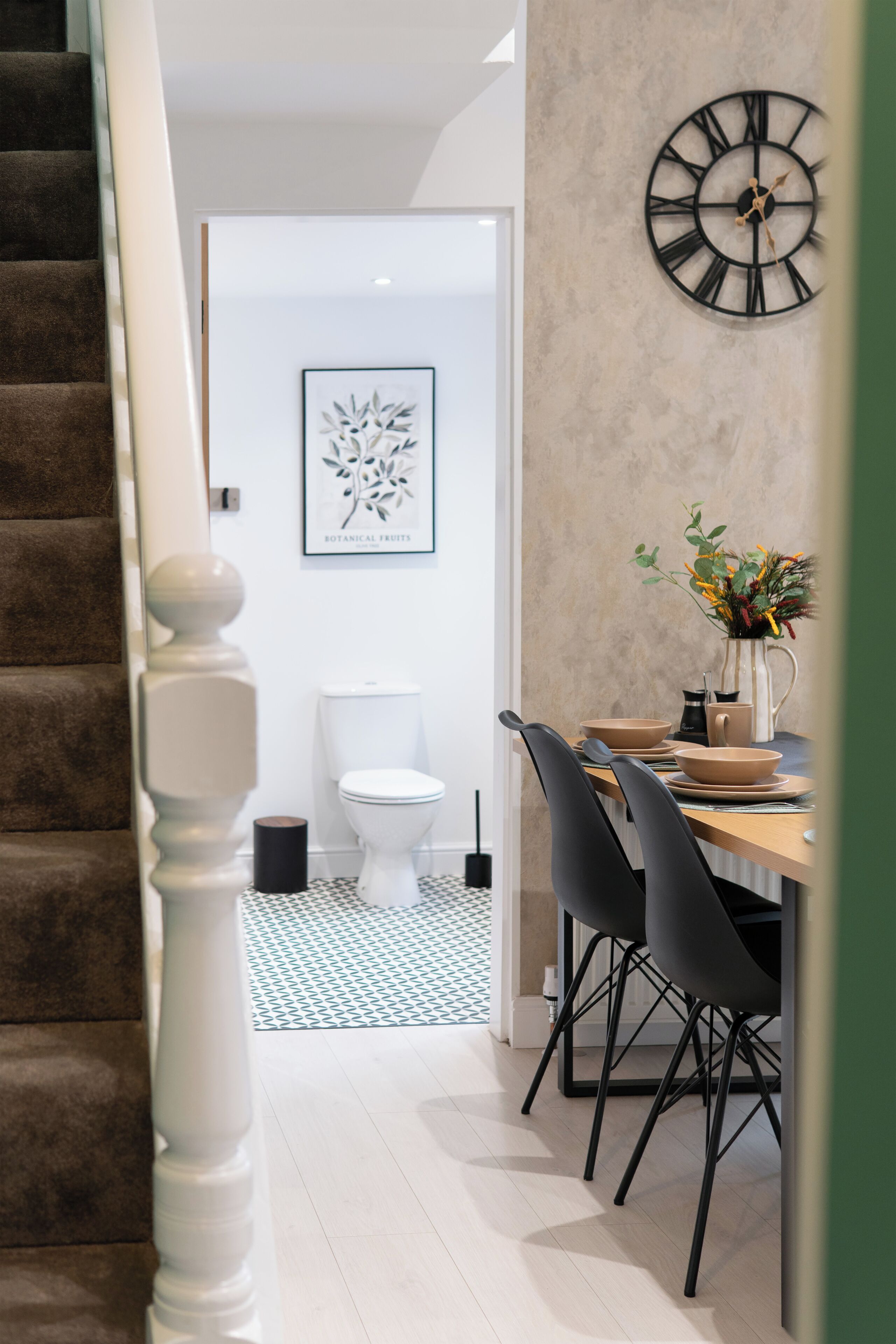 Un interior moderno con una escalera, una zona de comedor con sillas y una mesa, un reloj en la pared y un inodoro.