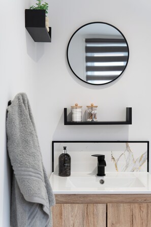 Un bany amb un lavabo blanc, un aixeta negre i un mirall a la part superior. Hi ha una tovallola penjada a la paret i una prestatgeria amb pots.