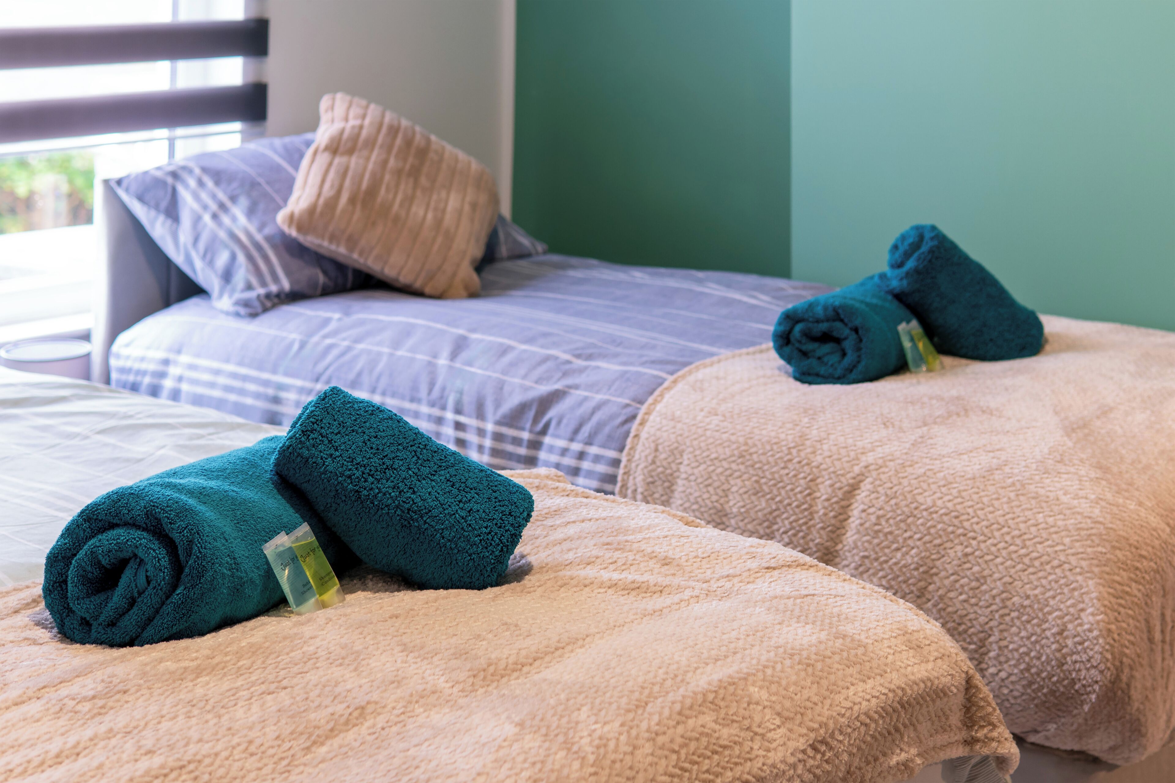 Ein Hotelzimmer mit zwei Betten, jedes mit einem gerollten Handtuch und einer Lotionflasche.