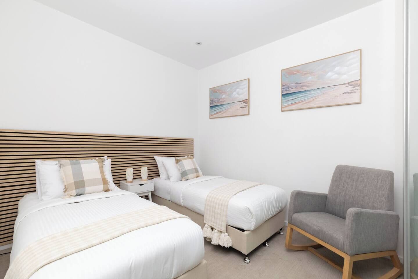 A bedroom with two beds, a nightstand, a chair, and two wall-mounted paintings.