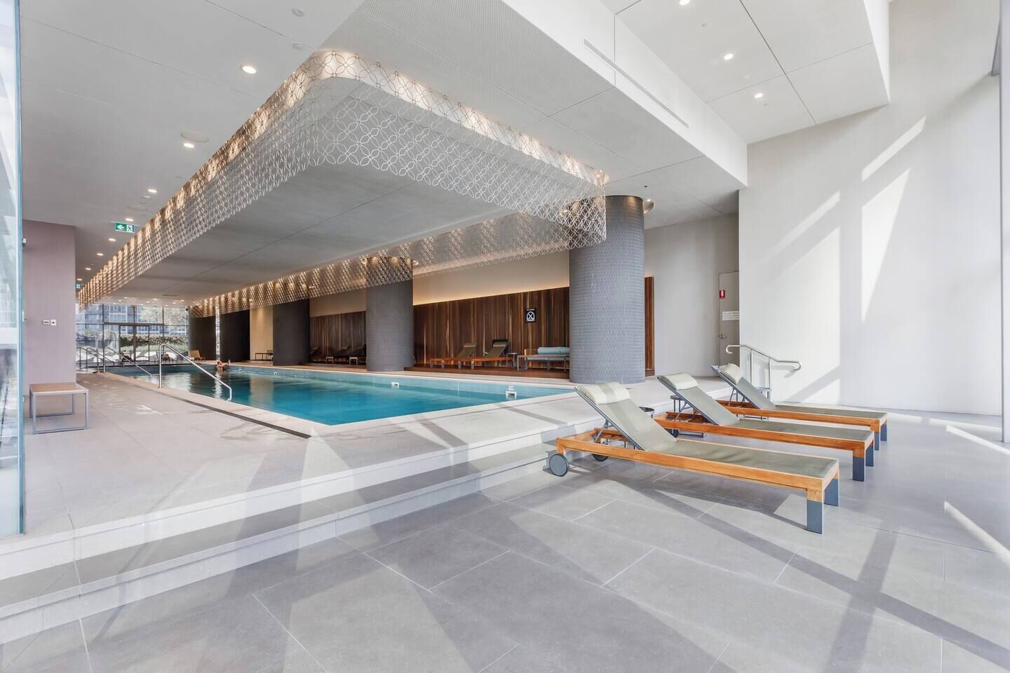 A modern indoor swimming pool with lounge chairs and a patterned ceiling.