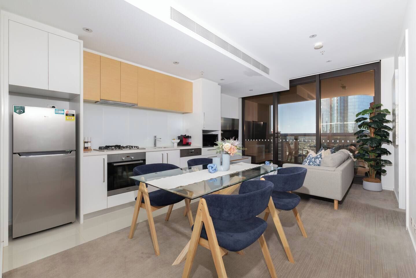 A modern kitchen with a built-in refrigerator, a dining area with a glass table and blue chairs, and a living area with a sofa and a large window overlooking a cityscape.
