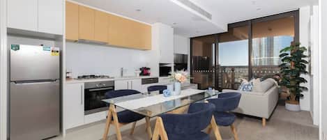 A modern kitchen with a built-in refrigerator, a dining area with a glass table and blue chairs, and a living area with a sofa and a large window overlooking a cityscape.
