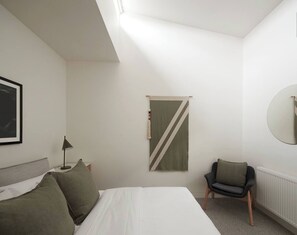 A bedroom with a bed, a chair, a bedside table, a lamp, a wall-mounted mirror, and a diagonal patterned wall hanging. - The Obscura Stylist Retreat near Cultural Hotspots (Richmond)