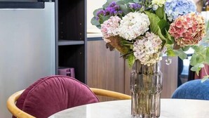 Une table ronde surmontée d’un vase de fleurs, une chaise en bois avec un coussin, et un coin cuisine à l’arrière-plan.