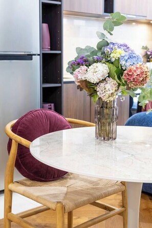 Ein runder Tisch mit Blumen in einer Vase, ein Holzhocker mit Kissen und im Hintergrund eine Küchenzeile.
