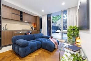 A modern living space with a sectional sofa, a glass coffee table, and a kitchen area with wooden cabinets. - Toorak Boutique Refined Living by Dining and Fashion (South Yarra)